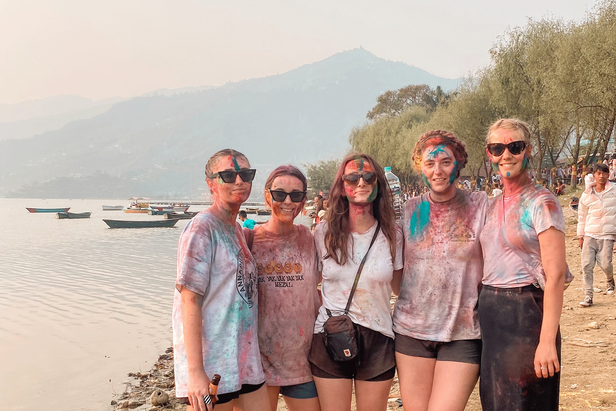 Group of girls in colourful clothes and with paint on their faces standing next to a lake in Pokhara, Nepal, with mountains in the background